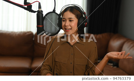 Charming Asian female podcaster wearing headphone and speaking into a microphone to recording podcast in home studio Charming Asian female podcaster wearing headphone and speaking into a microphone to recording podcast in home studio 94763379