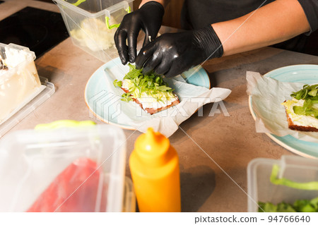 Cook hands preparing and making sandwich. Preparing healthy vegetarian bruschettas in dark gloves. Sandwich with soft cheese, arugula and cucumber. Cook hands preparing and making sandwich. Preparing healthy vegetarian bruschettas in dark gloves. Sandwich with soft cheese, arugula and cucumber. 94766640