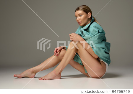 Portrait of young stylish woman sitting on floor in underwear and mint trench coat isolated over dark grey background Portrait of young stylish woman sitting on floor in underwear and mint trench coat isolated over dark grey background 94766749