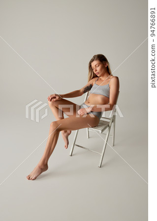 Portrait of young slim woman sitting on chair, posing in cotton underwear isolated over grey studio background Portrait of young slim woman sitting on chair, posing in cotton underwear isolated over grey studio background 94766891