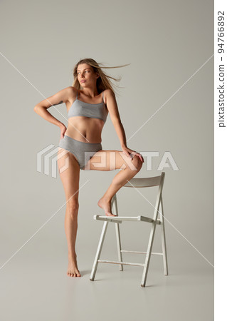 Portrait of slender young woman in cotton underwear leaning on chair isolated over grey studio background. Comfortable lingerie Portrait of slender young woman in cotton underwear leaning on chair isolated over grey studio background. Comfortable lingerie 94766892