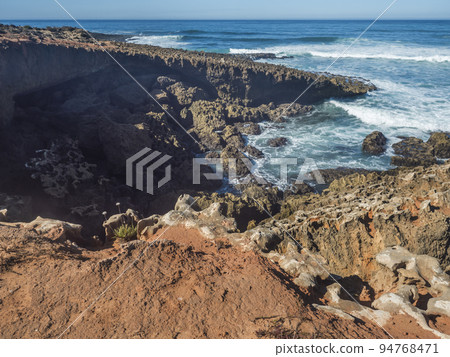 View of wild Rota Vicentina coast with ocean waves and red and orange sharp rock formations near Vila Nova de Milfontes, Portugal. Sunny day, blue sky 94768471
