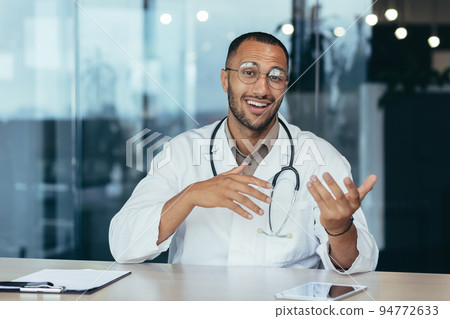 Webcam view, cheerful and successful family doctor consults patients remotely online, video call of man in medical coat smiling and looking at camera, working inside clinic office. Webcam view, cheerful and successful family doctor consults patients remotely online, video call of man in medical coat smiling and looking at camera, working inside clinic office. 94772633