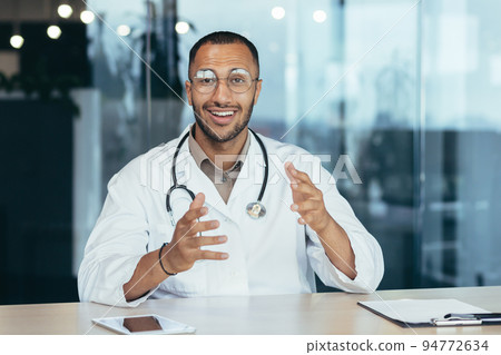 Webcam view, cheerful and successful family doctor consults patients remotely online, video call of man in medical coat smiling and looking at camera, working inside clinic office. Webcam view, cheerful and successful family doctor consults patients remotely online, video call of man in medical coat smiling and looking at camera, working inside clinic office. 94772634