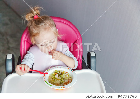 Infant baby with dirty face eating soup herself with spoon. Cute little girl sitting in high baby chair in kitchen at home. Maternity family childhood concept Infant baby with dirty face eating soup herself with spoon. Cute little girl sitting in high baby chair in kitchen at home. Maternity family childhood concept 94772769