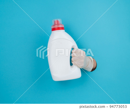 A woman is holding a white bottle of detergent. Woman's hand sticking out of blue paper background. A woman is holding a white bottle of detergent. Woman's hand sticking out of blue paper background. 94773053