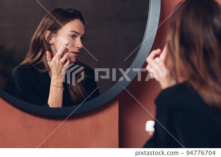 Young woman in front of the mirror putting cream on her face Young woman in front of the mirror putting cream on her face 94776704