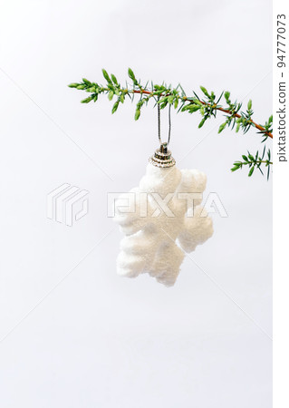 Happy holidays. Soft selective focus on white snowflake Christmas toy hanging on green branch of Christmas tree, juniper on white background. Minimal style. Happy holidays. Vertical Photo. Copy space 94777073