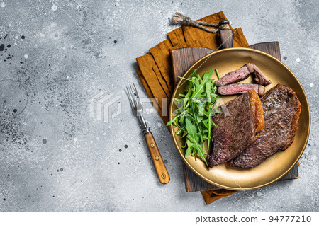 Roast picanha or top sirloin beef meat steak on a plate with arugula salad. Gray background. Top view. Copy space Roast picanha or top sirloin beef meat steak on a plate with arugula salad. Gray background. Top view. Copy space 94777210