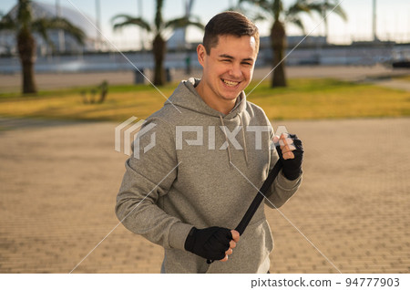 Caucasian man in hoodie bandaging his hands before boxing class outdoors. Caucasian man in hoodie bandaging his hands before boxing class outdoors. 94777903