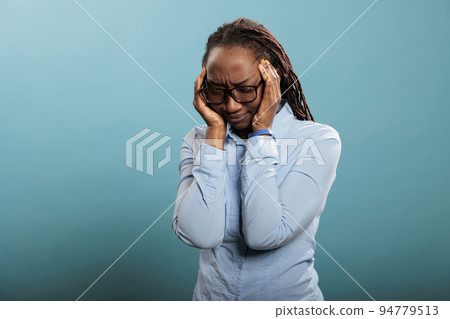 Overworked and stressed young adult person putting pressure on temples with fingers because of painful headache. African american woman suffering from migraine on blue background. Overworked and stressed young adult person putting pressure on temples with fingers because of painful headache. African american woman suffering from migraine on blue background. 94779513