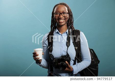 Smiling heartily young adult woman having DSLR device and cup of coffee standing on blue background. Confident and positive professional photographer posing for camera. Studio shot. 94779517