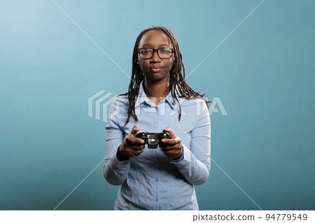 African american woman with modern console controller playing videogames on blue background. Young adult person enjoying competitive gaming while having gamepad. Studio shot. African american woman with modern console controller playing videogames on blue background. Young adult person enjoying competitive gaming while having gamepad. Studio shot. 94779549