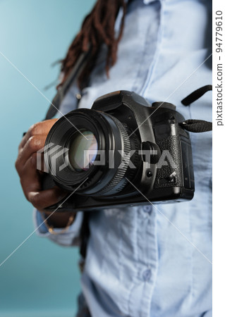Close up of young adult photograhpy entusiast holding a modern camera device while standing on blue background. Professional photographer with DSLR enjoying leisure time. Studio shot 94779610