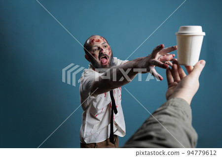 Spooky horror monster wanting cup of coffee, chasing after hand with carton cup with drink. Aggressive scary zombie eating brain and looking dangerous with bloody scars or wounds. 94779612