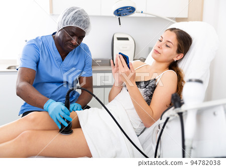 Woman using smartphone during cosmetological body procedure in clinic 94782038