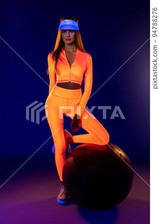 Stylish fit sportswoman in neon activewear standing in studio with fit ball under ultraviolet light 94788276