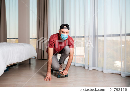 A young man in a medical mask poses on a longboard. Home interior. The concept of quarantine and activities 94792156