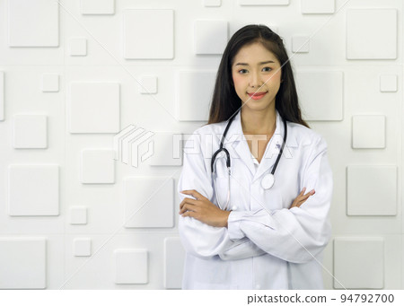 Young asian doctor in white gown and stethoscope stand with arm cross in front of white rectangle pattern ceramic wall. 94792700