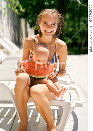 Mom sits on a sun lounger with a child and feeds him a watermelon 94793003