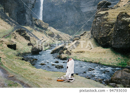 Man and woman in a blanket stand on the banks of a river in the mountains. Iceland 94793030