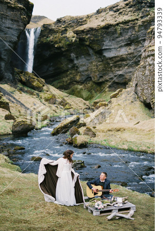 Groom plays the guitar next to the bride standing in a blanket on the river bank Groom plays the guitar next to the bride standing in a blanket on the river bank 94793339