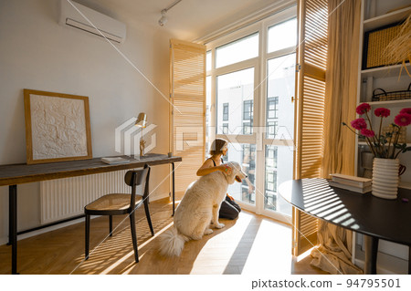 Woman stands with dog by the window blinds at home 94795501