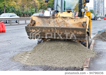A wide bucket of a road excavator unloads a mixture of sand gravel onto the asphalt for road repair. A wide bucket of a road excavator unloads a mixture of sand gravel onto the asphalt for road repair. 94796036