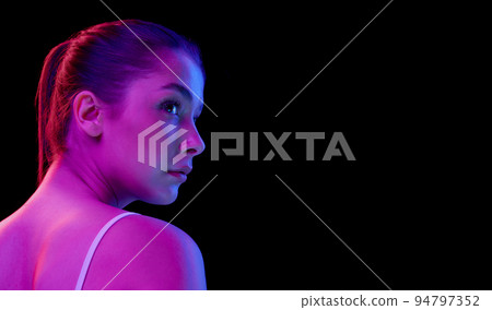 Cose-up portrait of young beautiful woman with clear spotless face looking away, posing isolated over black background in neon light. Side view Cose-up portrait of young beautiful woman with clear spotless face looking away, posing isolated over black background in neon light. Side view 94797352