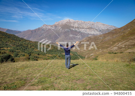 An adult man stands on top of a mountain with outstretched arms. 94800455