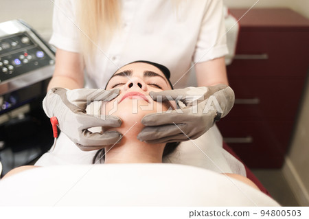 Doctor cosmetologist does facial massage to the patient with electric gloves.. Doctor cosmetologist does facial massage to the patient with electric gloves.. 94800503