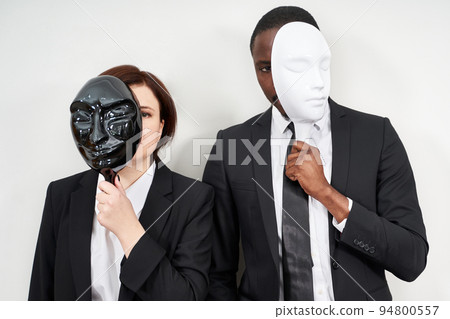 African man and Caucasian woman wearing black suits hiding face with plastic masks 94800557