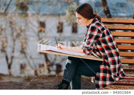 Young woman artist draws a picture while sitting on a bench. Painter artist holds a wooden tablet for painting and a brush in his hand 94801984