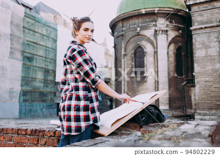 Woman is painting directly on the city street against the background of old architecture. Young painter artist in work. Woman is painting directly on the city street against the background of old architecture. Young painter artist in work. 94802229