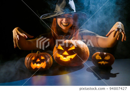 An ominous witch in a hat conjures over a jack-o-lantern. Traditional halloween characters. Mystical fog creeps over pumpkins with carved terrible faces. Woman in carnival costume. 94807427
