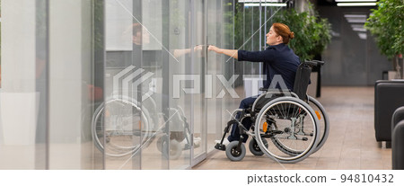Red-haired caucasian woman in a wheelchair trying to open the door in the office.  94810432