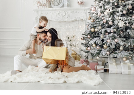Mother, father and two their sons sitting near Christmas tree. Excited family opening Christmas gifts at home. Woman, man and two boys wearing white clothes in white room. 94813953