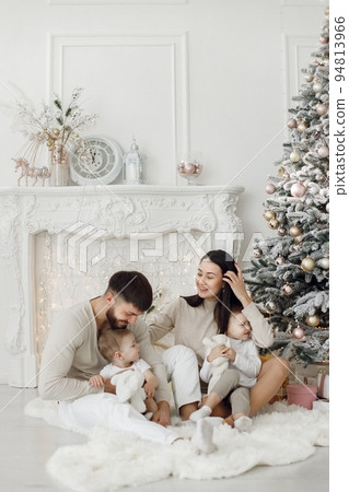 Mother, father and two their sons sitting near Christmas tree. Excited family opening Christmas gifts at home. Woman, man and two boys wearing white clothes in white studio. 94813966