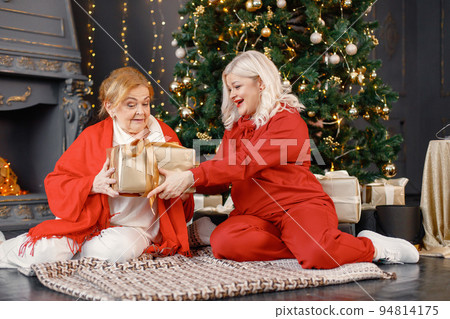 Two women celebrating Christmas together. Women sitting near Christmas tree with gift box and posing for a photo. Red-haired old woman and her adult blonde daughter wearing red clothes. Two women celebrating Christmas together. Women sitting near Christmas tree with gift box and posing for a photo. Red-haired old woman and her adult blonde daughter wearing red clothes. 94814175