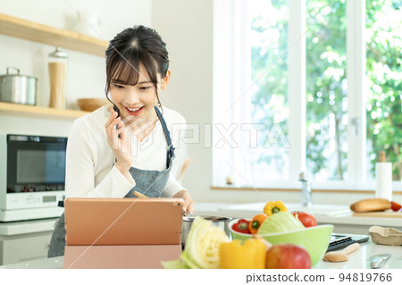 A young woman wearing an apron cooking while looking at a recipe A young woman wearing an apron cooking while looking at a recipe 94819766