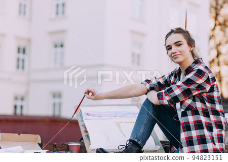 Woman artist paints a picture while sitting on a brick wall on a background of old architecture. Girl spends leisure drawing pictures on a city street.. 94823151