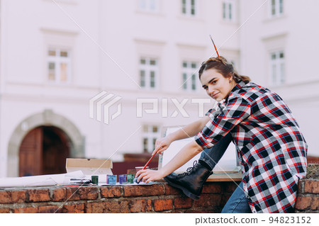 Woman artist paints a picture while sitting on a brick wall on a background of old architecture. Girl spends leisure drawing pictures on a city street.. Woman artist paints a picture while sitting on a brick wall on a background of old architecture. Girl spends leisure drawing pictures on a city street.. 94823152