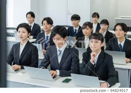 Young men and women listening to a seminar while looking at a large monitor Young men and women listening to a seminar while looking at a large monitor 94824017