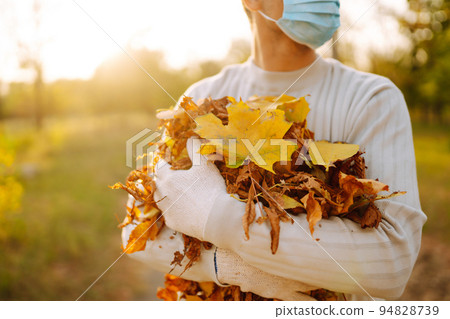Man in protective medical mask cleans autumn leaves in the park. Man in gloves collects. 94828739
