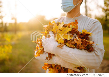Man in protective medical mask cleans autumn leaves in the park. Man in gloves collects. 94828759