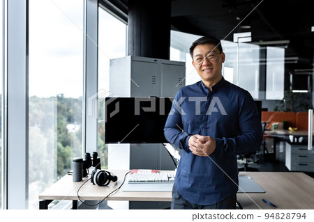 Programmer in glasses and shirt smiling and looking at camera, Asian man working inside modern computer company, working in office with several computers and monitors. 94828794