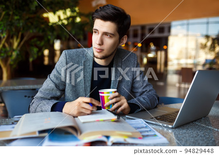 Shot of young male student sitting at table and writing on notebook. Male student studying in cafe. Shot of young male student sitting at table and writing on notebook. Male student studying in cafe. 94829448