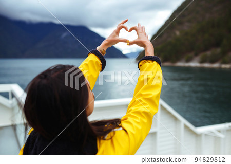 The girl tourist holding heart shaped hands and looking on the majestic mountains. The girl tourist holding heart shaped hands and looking on the majestic mountains. 94829812
