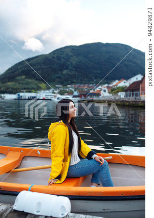 Girl tourist in yellow jacket is sitting and posing in a boat against the backdrop of the mountains 94829871