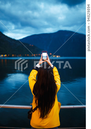 The girl tourist takes a photo on the phone by the lake in Norway. Active woman relaxing . The girl tourist takes a photo on the phone by the lake in Norway. Active woman relaxing . 94829894